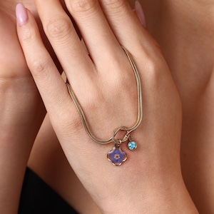 May include: A gold-toned double-strand chain bracelet featuring a knot detail. The bracelet includes two charms: one is a four-petaled purple flower encased in clear resin, and the other is a small, round, bezel-set bright blue gemstone.