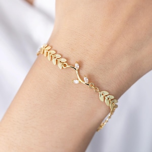 May include: A gold bracelet featuring a delicate leaf design with clear crystal accents. The bracelet is made of a chain with small leaves and a larger leaf design with a crystal in the centre.