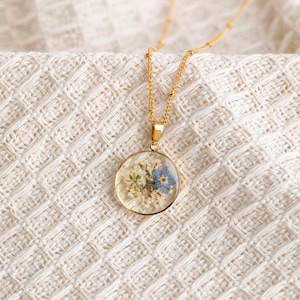 May include: A gold chain necklace with a round pendant featuring pressed blue and white flowers encased in clear resin.