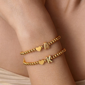 May include: Two gold-toned beaded bracelets, each featuring a small polished heart charm and a single letter charm. The top bracelet displays the letter A, while the bottom bracelet displays the letter K. The beads are uniform and spherical in shape.