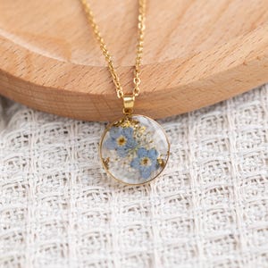 May include: A gold chain necklace with a round pendant featuring a pressed blue flower design encased in clear resin. The pendant has a gold flecked background.