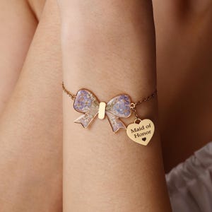 May include: A gold-toned chain bracelet featuring a bow-shaped charm filled with pressed purple and white flowers. A small, heart-shaped gold-toned pendant hangs from the chain, engraved with the text "Maid of Honor" and a tiny heart symbol.