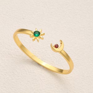 Può includere: Un anello aperto color oro caratterizzato da un motivo a sole con una gemma verde centrale su un'estremità e un motivo a mezzaluna con una gemma rossa centrale sull'altra. La fascia è liscia e circolare con un'apertura regolabile.