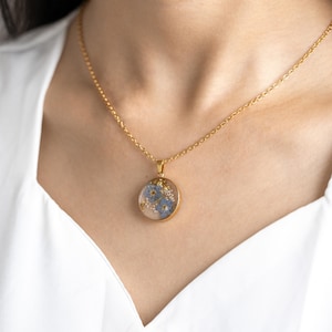 May include: A gold chain necklace with a round pendant featuring a delicate arrangement of blue forget-me-not flowers pressed inside clear resin.