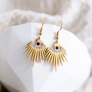 May include: Gold tone earrings with a sunburst design and a blue evil eye charm. The earrings are adorned with clear rhinestones.