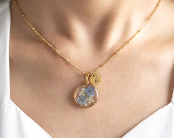 May include: A gold chain necklace with a round pendant featuring pressed blue and white flowers encased in clear resin. A smaller gold disc pendant with an engraved letter 'L' hangs above the floral pendant.
