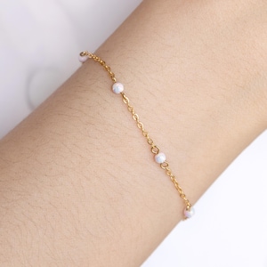 Op de afbeelding: Een delicate goudkleurige kettingarmband versierd met kleine, iriserende witte kralen. De armband wordt om een pols gedragen en toont zijn eenvoudige maar elegante ontwerp. De kralen zijn gelijkmatig over de ketting verdeeld.