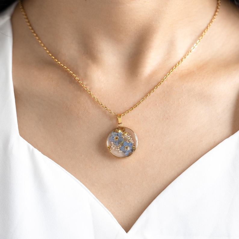 May include: A gold chain necklace with a round pendant featuring pressed blue flowers encased in clear resin.