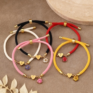 May include: A collection of five adjustable cord bracelets in black, white, pink, red, and yellow. Each bracelet features gold-toned metal sliders, a heart-shaped charm engraved with a letter, and a small circular pendant containing a pressed flower.