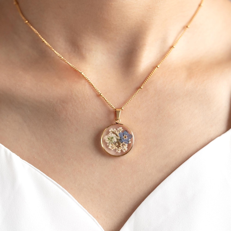 May include: A gold chain necklace with a round pendant featuring pressed flowers in shades of blue, white, and green.