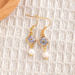 May include: A pair of gold-coloured drop earrings. Each earring features a four-leaf clover-shaped frame with a light blue flower encased within. A white pearl hangs below each frame, creating a delicate and elegant design.