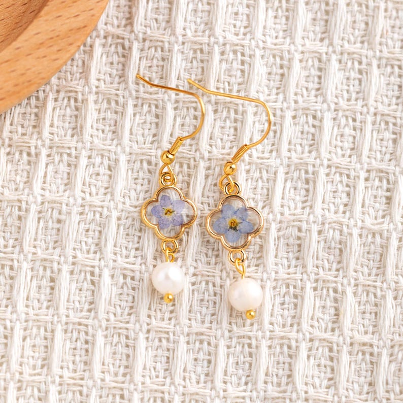 May include: A pair of gold-coloured drop earrings. Each earring features a four-leaf clover-shaped frame with a light blue flower encased within. A white pearl hangs below each frame, creating a delicate and elegant design.