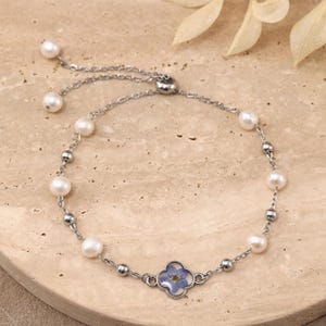 May include: A silver-toned chain bracelet featuring white pearls and small metallic beads. The bracelet includes a central four-leaf clover charm containing a pressed blue flower and an adjustable sliding clasp with two dangling pearls at the ends.