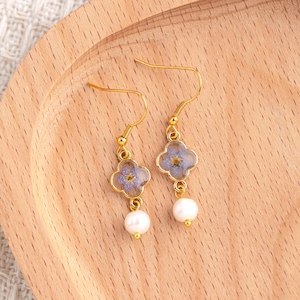 May include: Gold-coloured earrings with a four-leaf clover design, each holding a small, light purple flower in clear resin. White pearl drops hang below. The earrings are presented on a wooden surface.