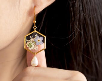 May include: A gold-toned hexagonal drop earring featuring clear resin encasing pressed white and light blue flowers. A small, white, teardrop-shaped pearl hangs from the bottom of the gold metal frame, suspended by a gold hook wire.