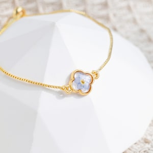 Pu&ograve; includere: Un delicato braccialetto a catena dorata con un ciondolo a forma di quadrifoglio con un fiore blu pressato.