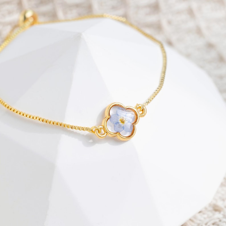 Pu&ograve; includere: Un delicato braccialetto a catena dorata con un ciondolo a forma di quadrifoglio con un fiore blu pressato.