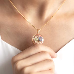May include: A gold chain necklace with a round pendant featuring pressed flowers in shades of blue, white, and green encased in clear resin.