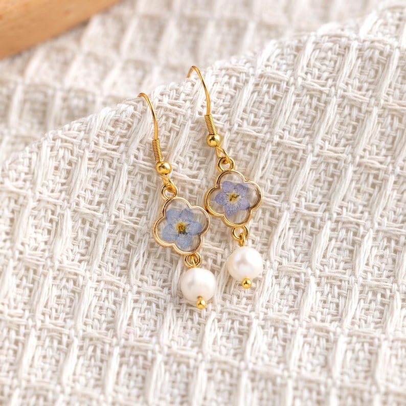 May include: Gold-tone drop earrings. Each earring has a four-leaf clover-shaped charm with a light blue flower set in clear resin. A white pearl hangs below each charm. The earrings are presented on a white textured surface.