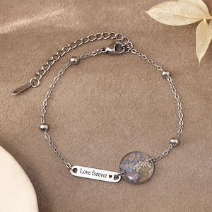 May include: A silver-toned metal chain bracelet featuring a small rectangular charm engraved with the text "Love forever" followed by a heart symbol, and a circular pendant containing pressed blue and white flowers encased in a clear resin.