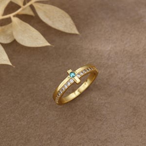 Peut inclure: Une bague dorée présentant un motif central en forme de croix orné d'une petite pierre précieuse ronde de couleur sarcelle. L'anneau est agrémenté d'une rangée de pierres transparentes et scintillantes serties pavé le long de la tige.