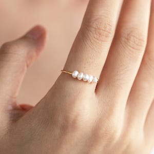 May include: A slender gold ring adorned with five small, white pearls. The ring's design is minimalist, with the pearls arranged in a line. The ring is displayed on a finger, showcasing its understated elegance. The pearls are approximately 0.3 cm in diameter.