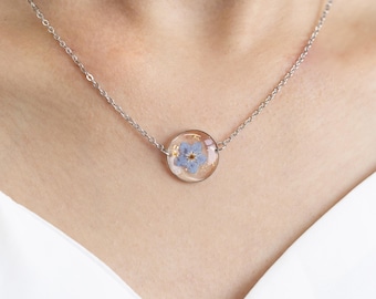 May include: A silver chain necklace with a round pendant featuring a preserved blue forget-me-not flower encased in clear resin.