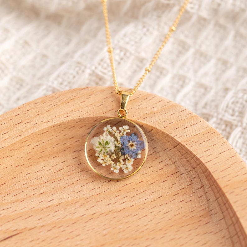 May include: A gold chain necklace with a round pendant featuring pressed blue and white flowers encased in clear resin.