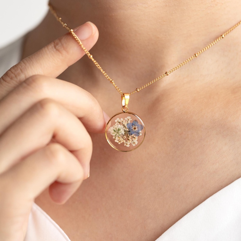 May include: A gold chain necklace with a round pendant featuring pressed blue and white flowers encased in clear resin.