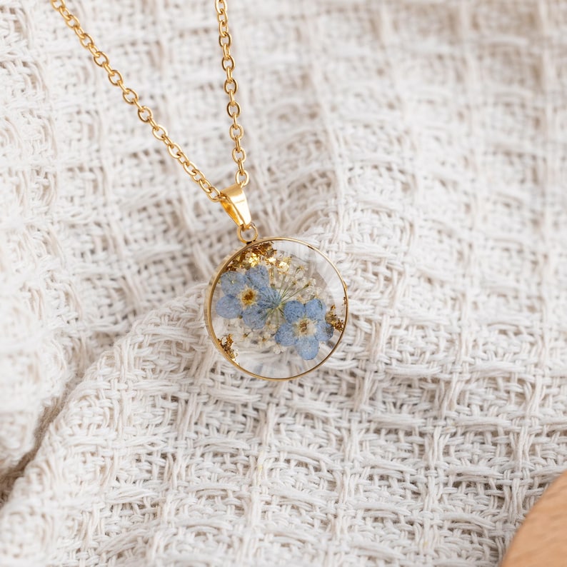 May include: A gold chain necklace with a round pendant featuring pressed blue flowers and gold flecks encased in clear resin.