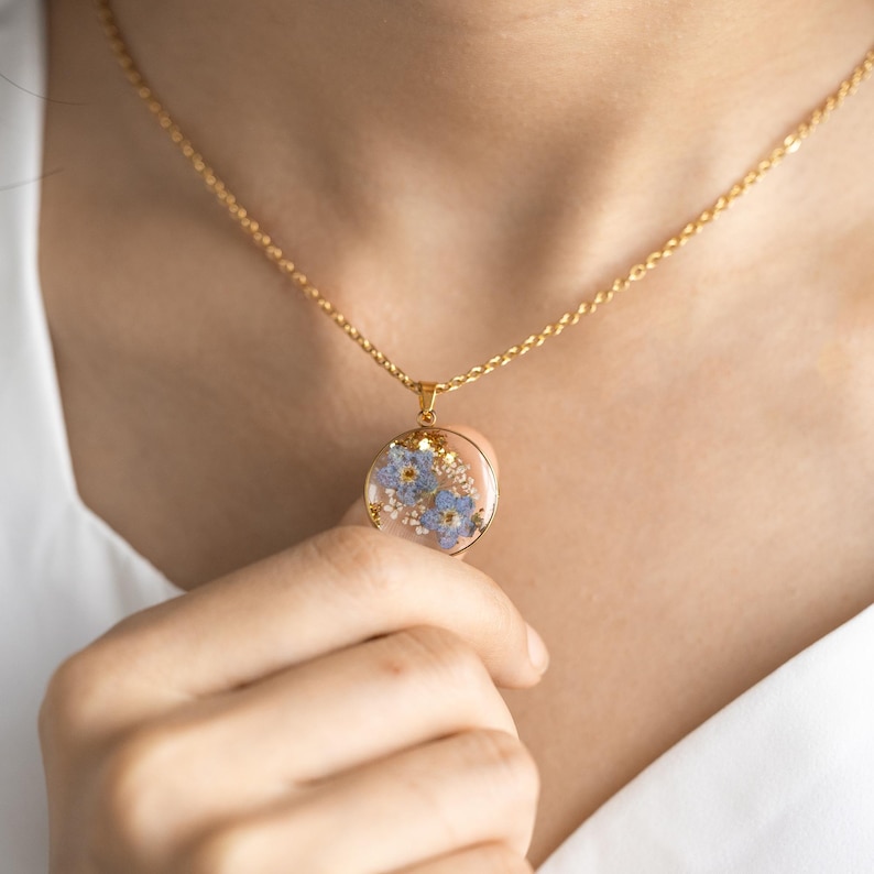 May include: A gold chain necklace with a round pendant featuring pressed blue forget-me-not flowers encased in clear resin.