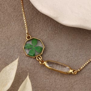 Puede incluir: Una pulsera de cadena dorada con dos dijes distintos. Un dije es un colgante hexagonal que contiene un trébol de cuatro hojas verde prensado dentro de resina transparente. El segundo dije es un colgante ovalado alargado con una superficie lisa y clara.