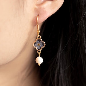 May include: Gold-coloured drop earring featuring a four-leaf clover design with a blue floral element and a small yellow centre. A white pearl hangs below the clover. The earring is shown against a person's ear and dark hair.
