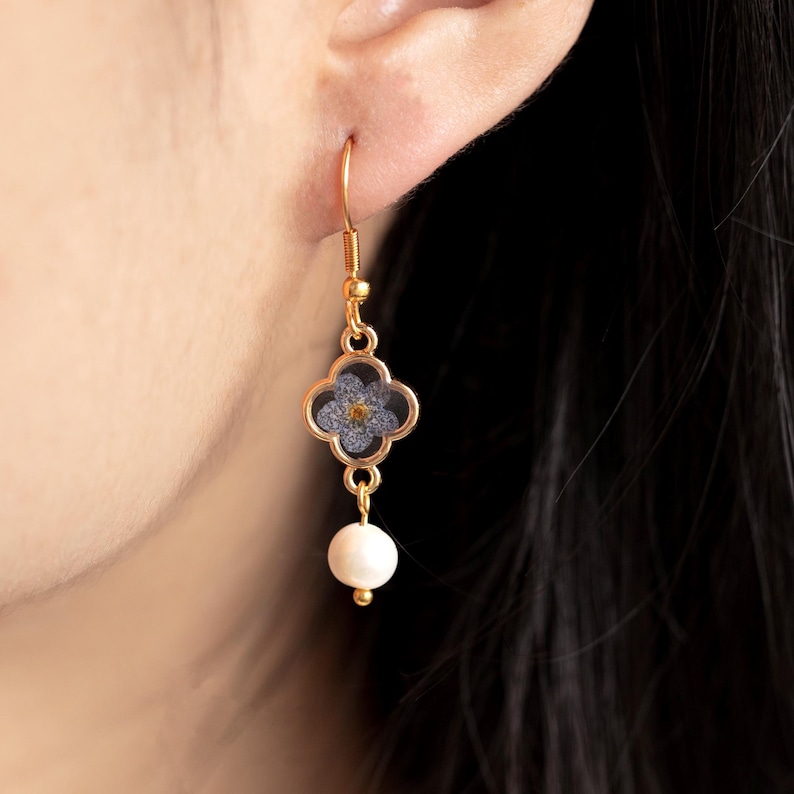 May include: Gold-coloured drop earring featuring a four-leaf clover design with a blue floral element and a small yellow centre. A white pearl hangs below the clover. The earring is shown against a person's ear and dark hair.