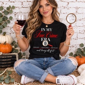May include: A black short-sleeve t-shirt featuring the white and red text IN MY True Crime ERA with a magnifying glass graphic over a fingerprint, plus the words PODCASTS, COLD CASES, RED FLAGS and and loving all of it! in white.