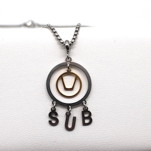 Könnte beinhalten: Eine silberfarbene Halskette mit einem Anhänger. Der Anhänger hat einen schwarzen Außenring, ein goldfarbenes Innendesign und die Buchstaben "SUB" darunter. Die Halskette wird auf einer weißen Oberfläche präsentiert.