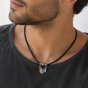 Könnte beinhalten: Eine schwarze Kordelkette mit einem kleinen Anhänger aus vertikalen, mehrfarbigen Stäben in Regenbogenfolge, akzentuiert durch eine silberfarbene Metallperle an der Oberseite des Anhängers, getragen zu einem dunkelgrauen T-Shirt.