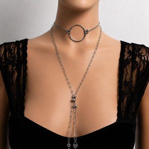 Può includere: Una collana girocollo argentata con un grande pendente circolare e una lunga collana a catena. La lunga catena presenta due piccole sfere argentate, un ciondolo a forma di chiave e un ciondolo a forma di lucchetto a cuore. Gioielli su manichino.