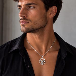 Op de afbeelding: Een zilverkleurige ronde hanger aan een ketting met een gedetailleerd silhouet van een hertenkop in het midden van een ring, hangend aan een bijpassende zilverkleurige ketting. De hanger bevat een klein hangslot-icoon in het frame.