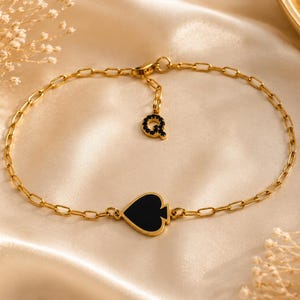 Könnte beinhalten: Ein goldfarbenes Kettenarmband mit einem zentralen schwarzen Anhänger in Pik-Form und einem kleinen, baumelnden Buchstaben-Anhänger Q, der mit winzigen schwarzen Steinen besetzt ist. Das Armband besteht aus länglichen ovalen Gliedern und einem Karabinerverschluss.