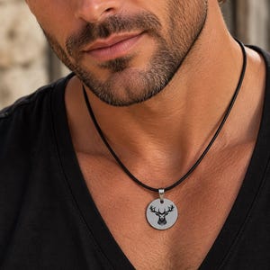 May include: A circular silver-toned metal pendant featuring a black engraved deer head silhouette with antlers. The pendant is suspended from a simple black cord necklace, resting against a plain black v-neck shirt.