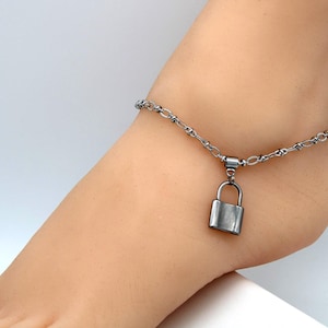 Könnte beinhalten: Eine silberfarbene Metall-Fußkette mit einer zarten Kette aus kleinen, sich wiederholenden ovalen Gliedern und einem einzelnen, glatten, rechteckigen Vorhängeschloss-Anhänger, der in der Mitte hängt. Die Oberfläche ist poliert.