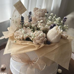 Könnte beinhalten: Eine dekorative Anordnung von Kerzen in Form von Blumen, einem Teddybären und kleinen Schokoriegeln, präsentiert in einer weißen runden Box mit einem Band. Die Anordnung umfasst Lavendel und Schleierkraut und erzeugt eine weiche, pastellfarbene Farbpalette.