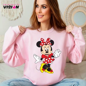 May include: A pink sweatshirt with a graphic of Minnie Mouse wearing a red polka dot dress and a red bow.