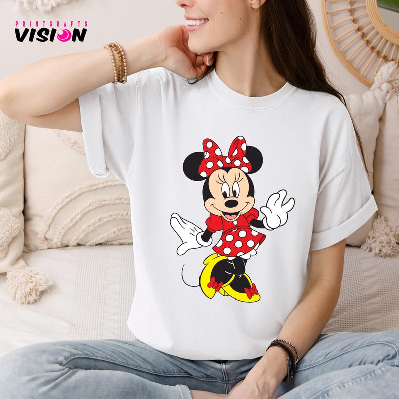 May include: White t-shirt with a red and white Minnie Mouse graphic. Minnie Mouse is wearing a red dress with white polka dots and a red bow with white polka dots. She is smiling and has her arms outstretched.