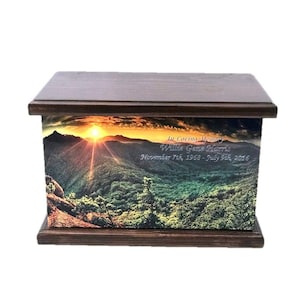 May include: A wooden cremation urn with a sunset over a mountain range scene. The urn has the text "In Loving Memory Willie Gene Hicks November 7th, 1953 - July 21st, 2016"