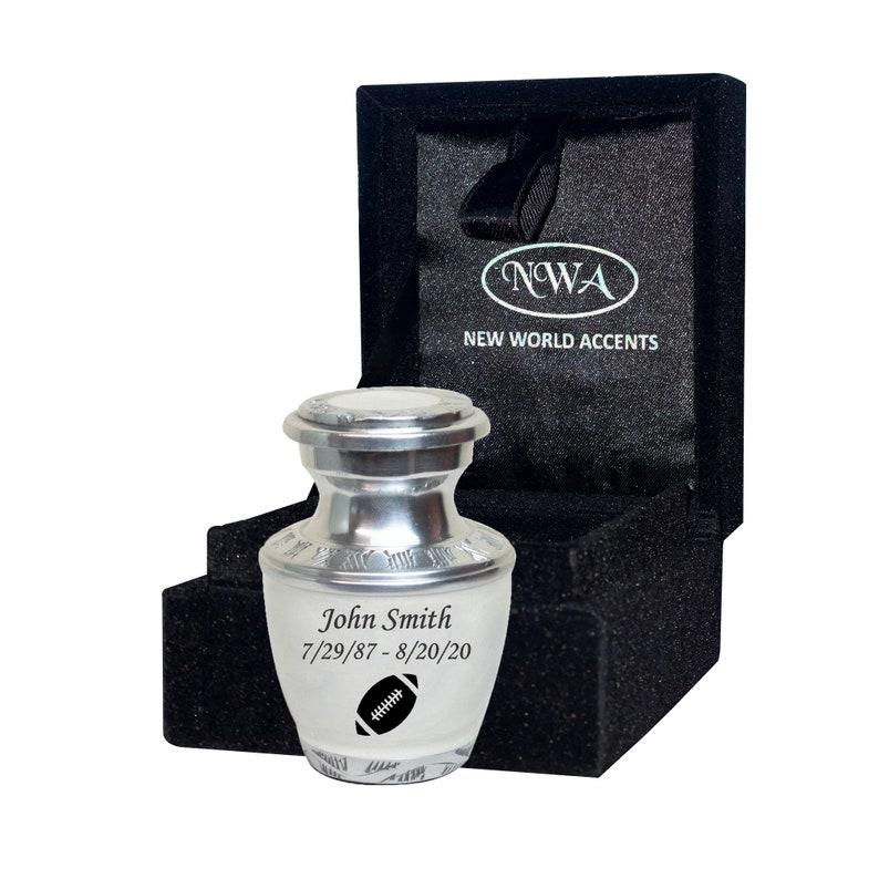 May include: A silver metal urn with a black football design and the inscription "John Smith 7/29/87 - 8/20/20". The urn is in a black velvet box with the inscription "NWA NEW WORLD ACCENTS".