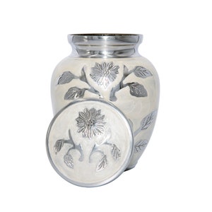 May include: White ceramic cremation urn with a silver floral design. The urn has a lid with a matching design.