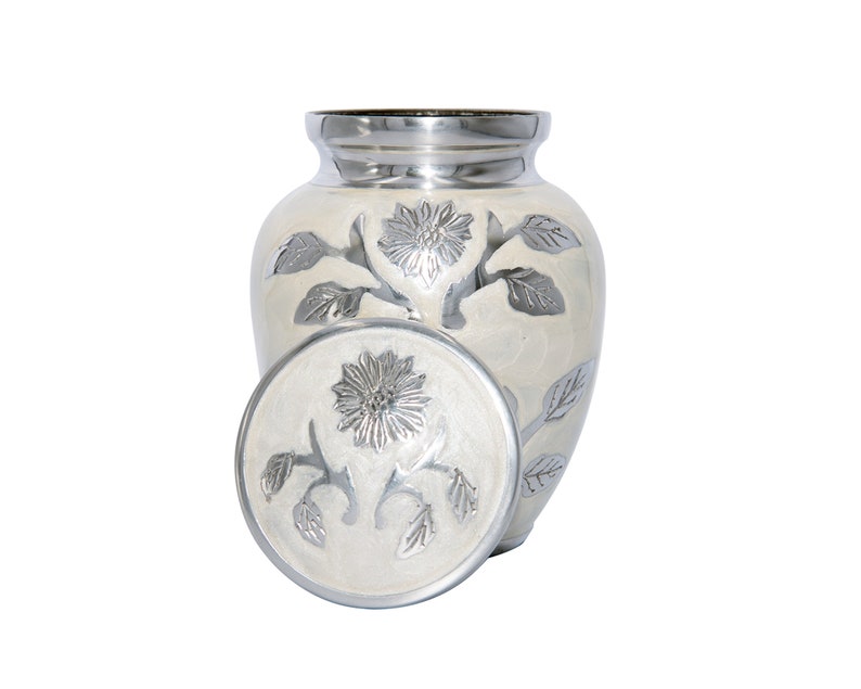 May include: White ceramic cremation urn with a silver floral design. The urn has a lid with a matching design.