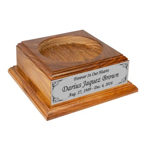 May include: A wooden urn with a square base and a circular opening. A silver plaque on the front reads "Forever In Our Hearts" and the name "Darius Jaquez Brown" with birth and death dates.